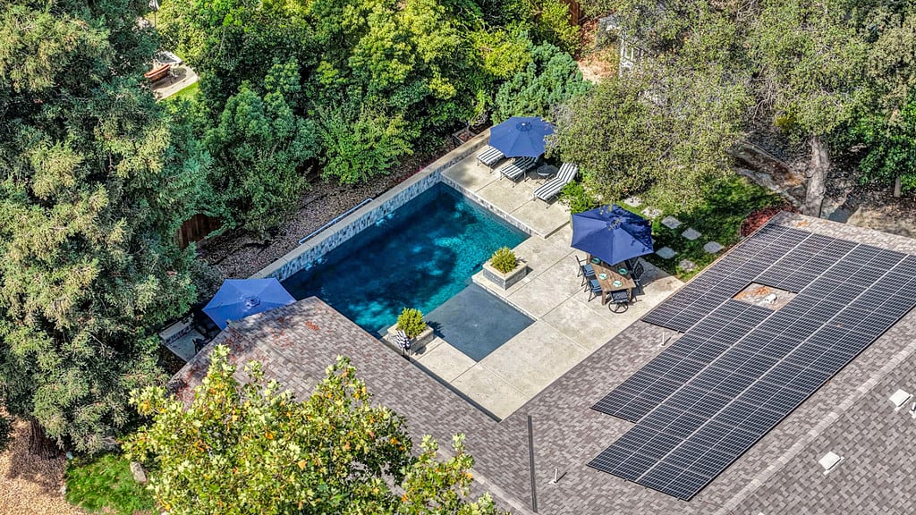 Aerial view of a backyard pool and patio beside a roof covered in solar panels.