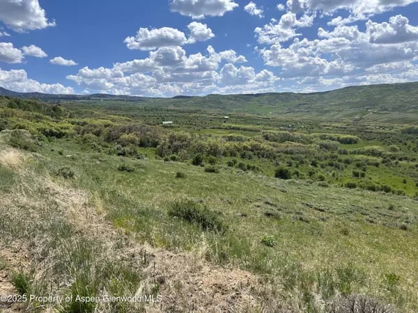 Rolling green hills and shrubland beneath a cloud-filled blue sky.