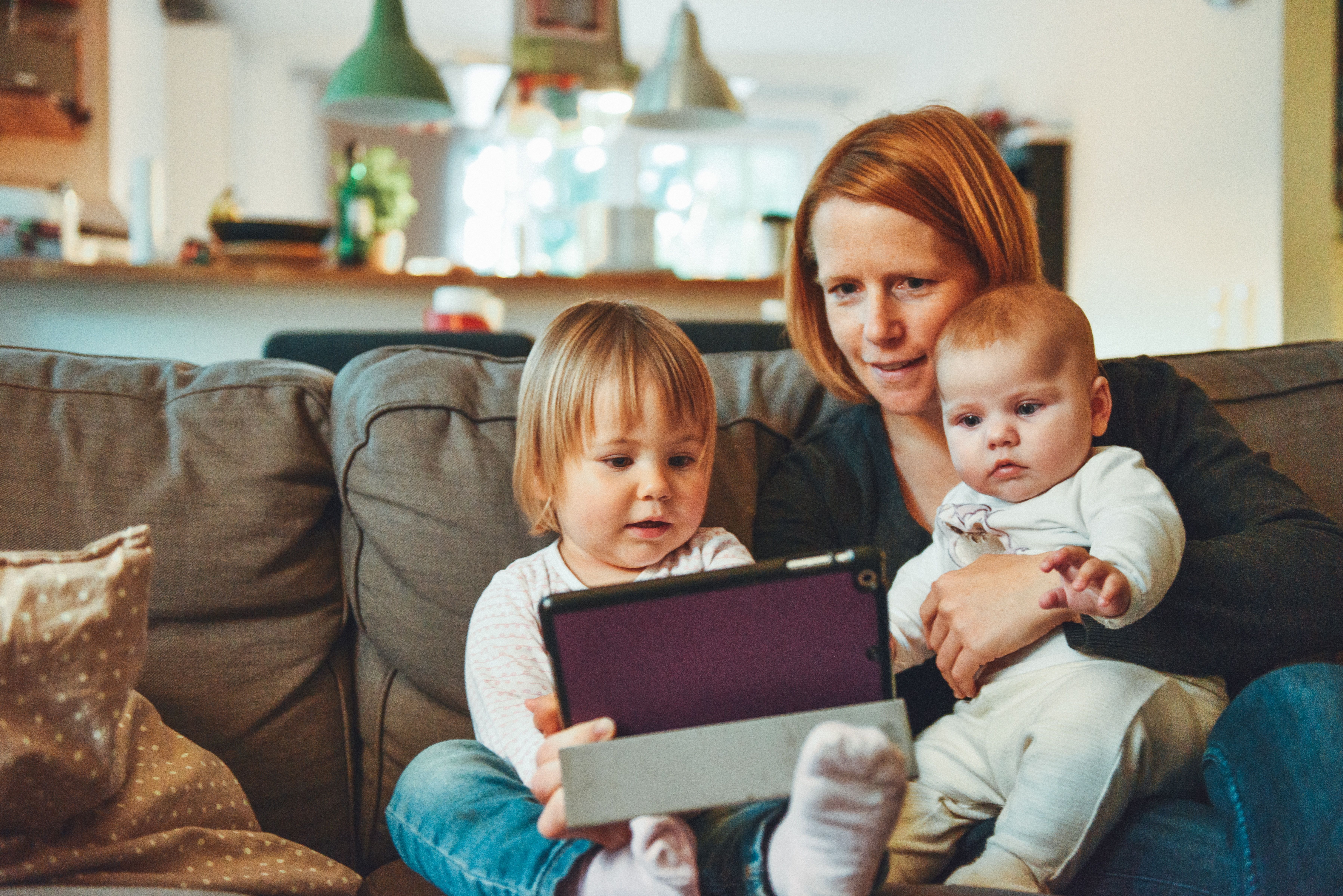 A woman sits with two young children on a sofa as they look at a tablet.