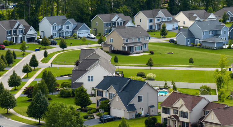 Suburban neighborhood view with rows of modern two-story houses in varied colors, surrounded by lush green lawns, trees, and a peaceful atmosphere.