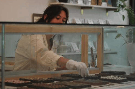 A gloved shop worker arranges chocolates in a glass display case.