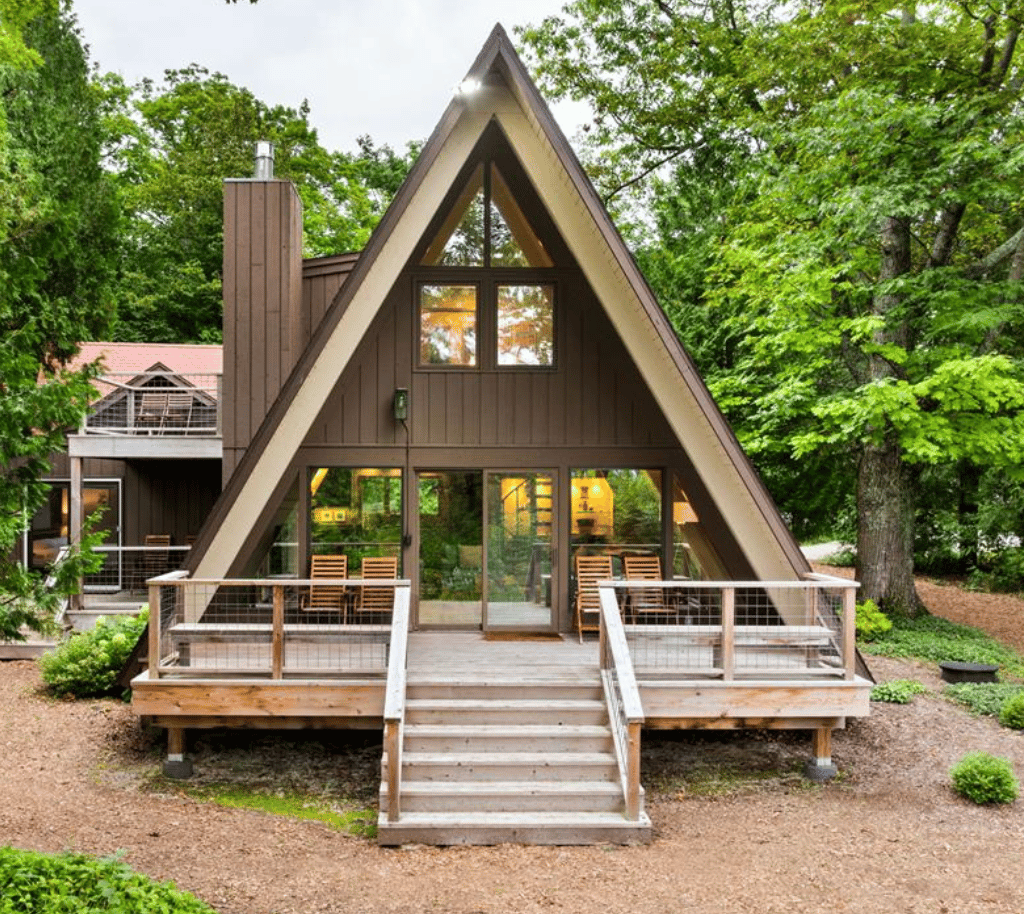 Wooded A-frame house with a raised wooden deck, outdoor chairs, and broad front windows.
