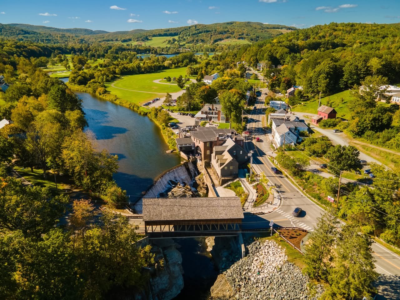 Vermont village and covered bridge spanning a river, surrounded by wooded hills and fields.