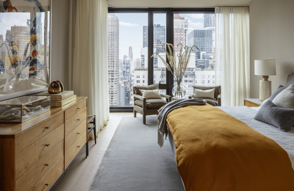 Furnished bedroom with a mustard-yellow bedspread and city views through large windows.