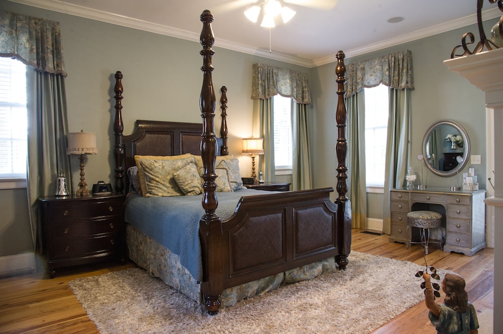 Master bedroom with a carved four-poster bed, blue bedding, curtained windows, and a dresser with a round mirror.