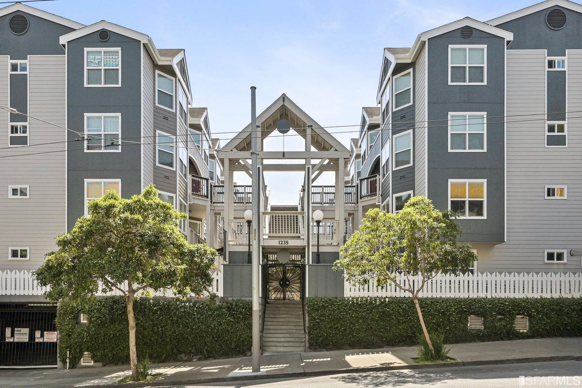 Multi-story residential building with a peaked central entrance, balconies, and trees along the sidewalk.