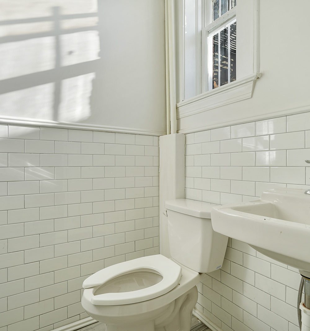 Bathroom with white subway tile, a toilet, and a wall-mounted sink beneath a high window.