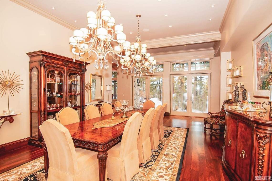 Formal dining room with ornate chandeliers, a carved wood table, cream chairs, and tall windows.