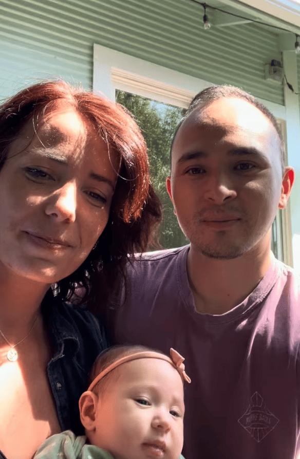 A woman and man pose with an infant on a sunlit porch.