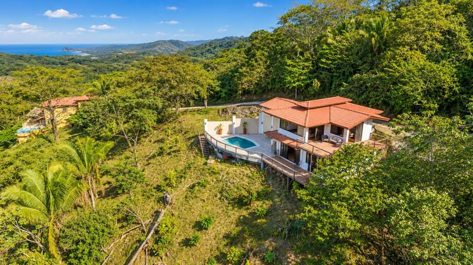 Aerial view of a red-tile-roof home with a pool amid tropical forest.