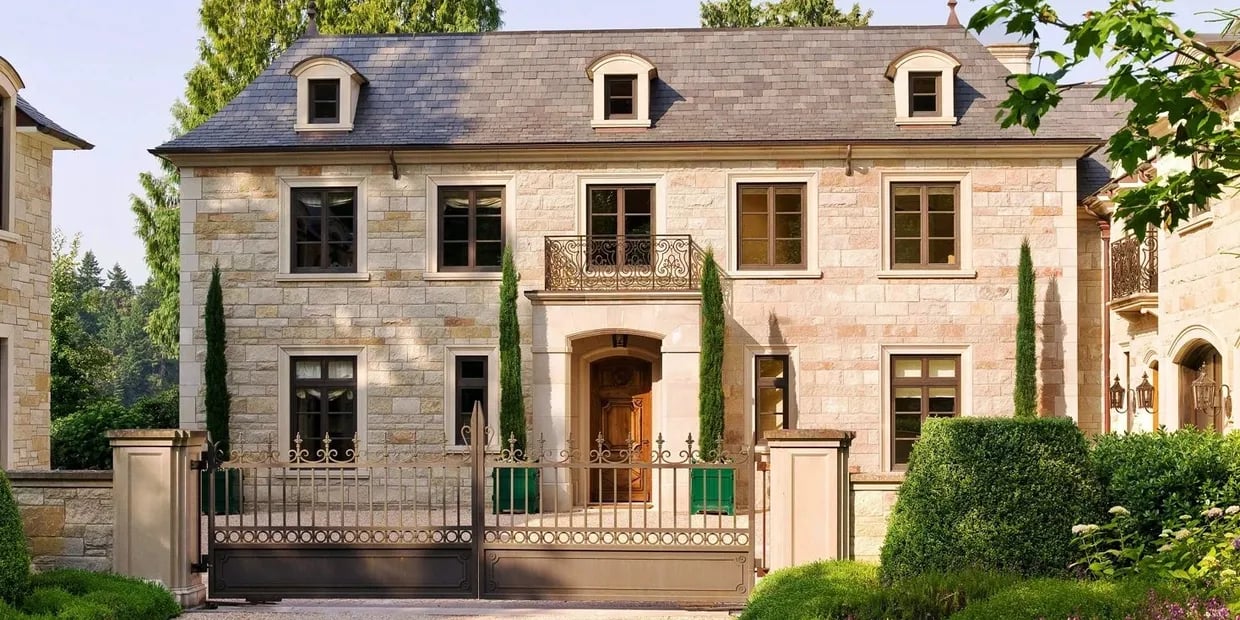 French country stone house with slate roof, dormer windows, wrought-iron balcony, and ornate front gate.
