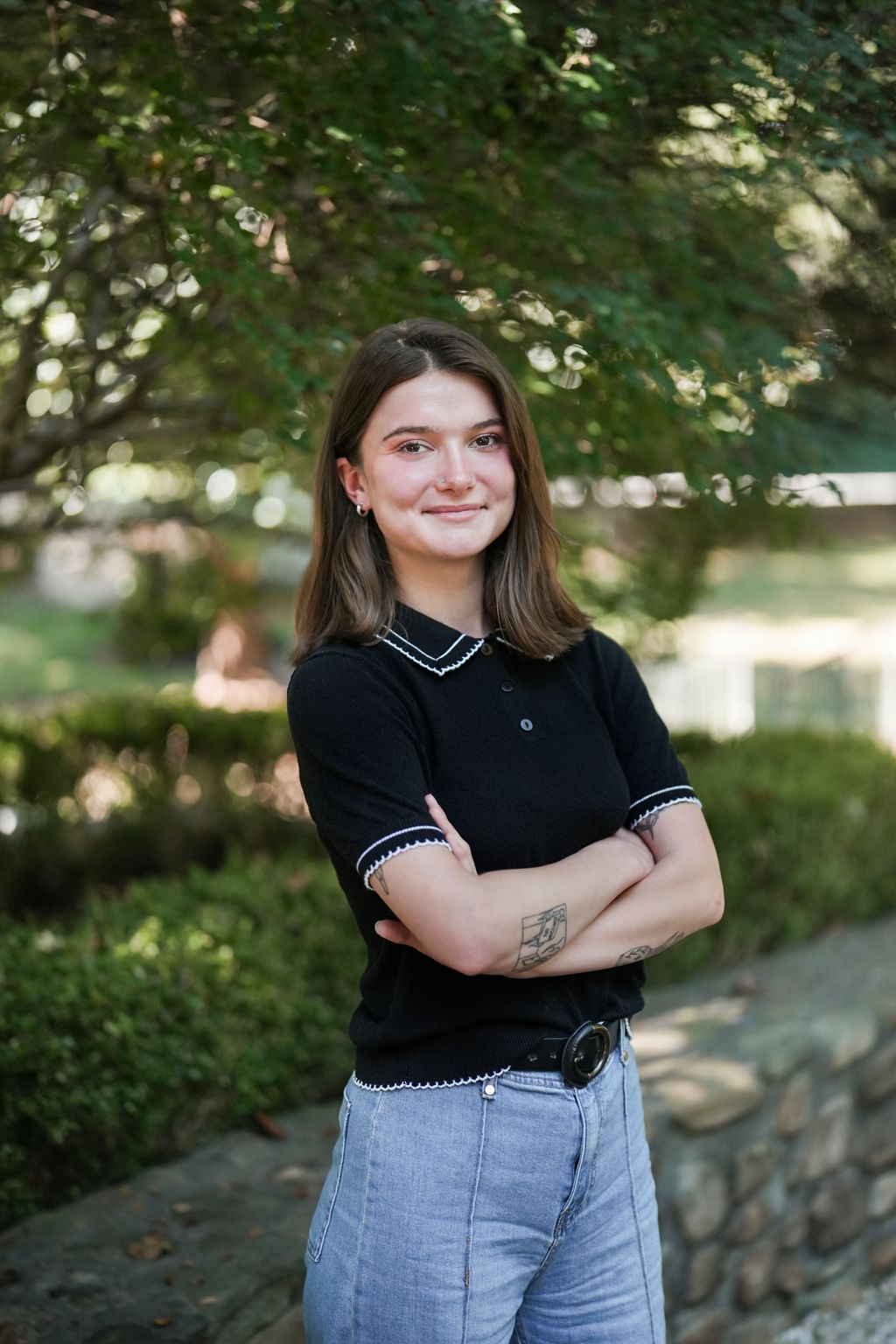 Teja Pope stands outdoors with folded arms, wearing a black top and light jeans.