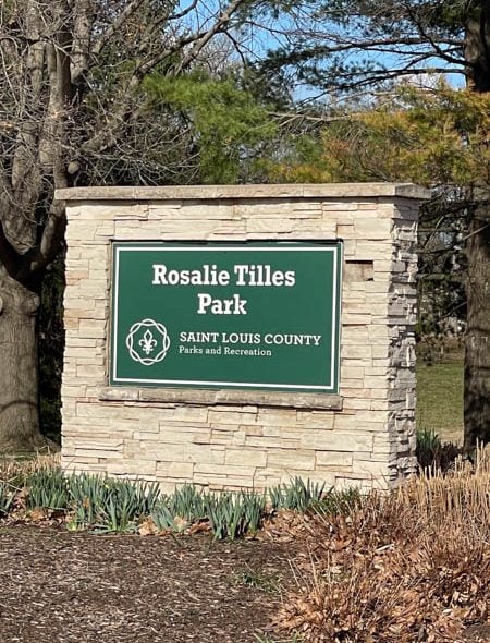 Rosalie Tilles Park sign mounted on a stone monument among trees and landscaping.