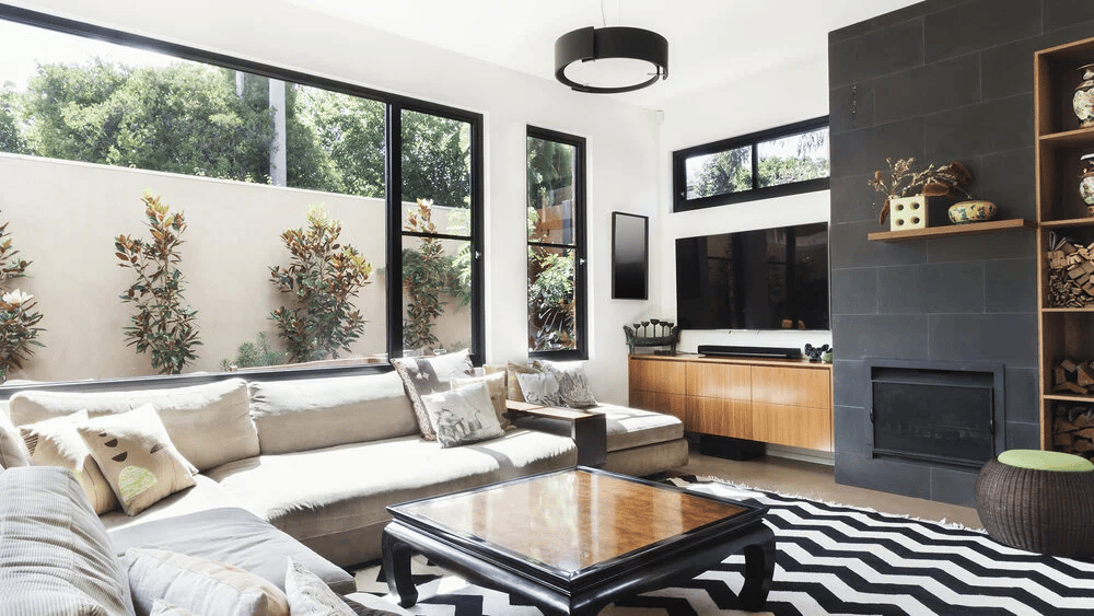 Modern living room with a cream sectional, large windows, geometric rug, and dark tiled fireplace wall.