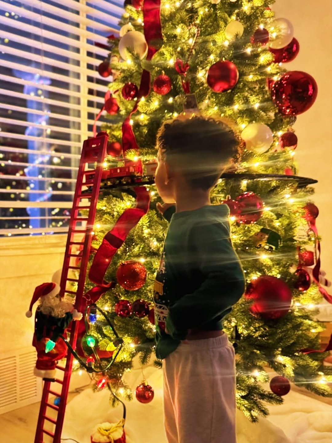 Child sitting near a glowing Christmas tree, gazing at twinkling holiday lights in a cozy living room.