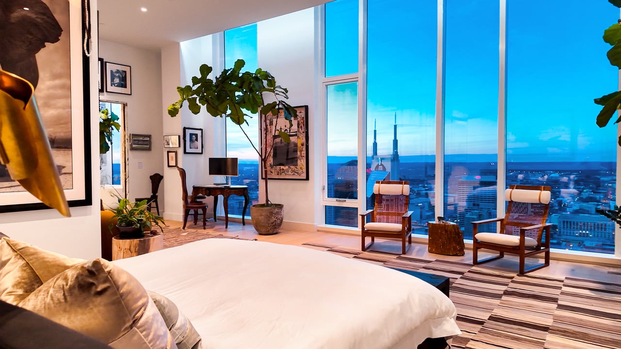 High-rise bedroom with floor-to-ceiling windows overlooking a city skyline at dusk.