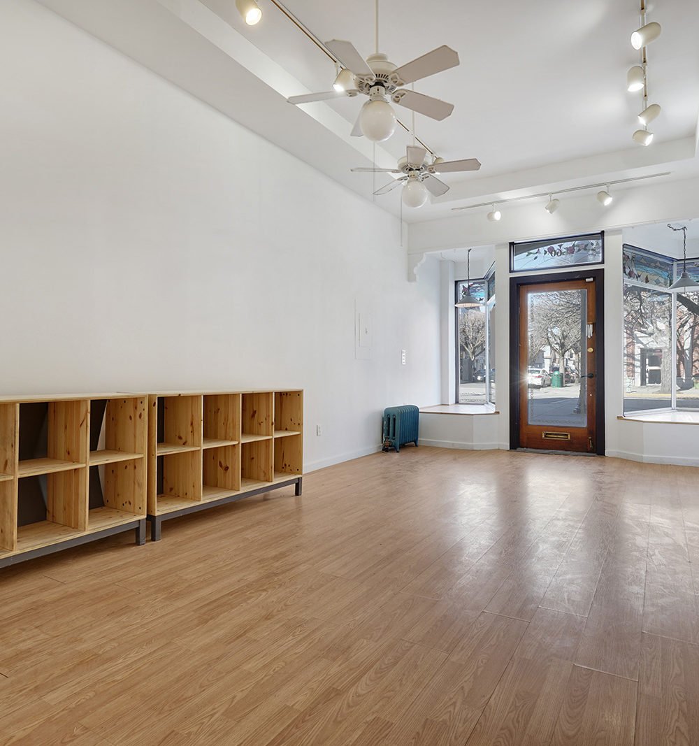 Vacant storefront with wooden shelving, plank flooring, ceiling fans, and a glass front entrance.