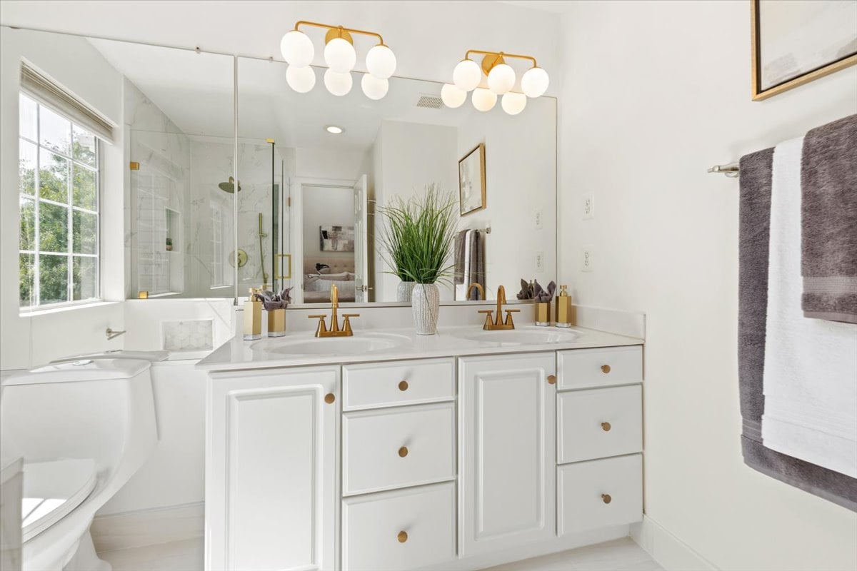 Bright bathroom with a white double vanity, gold fixtures, glass shower, and window beside the tub.