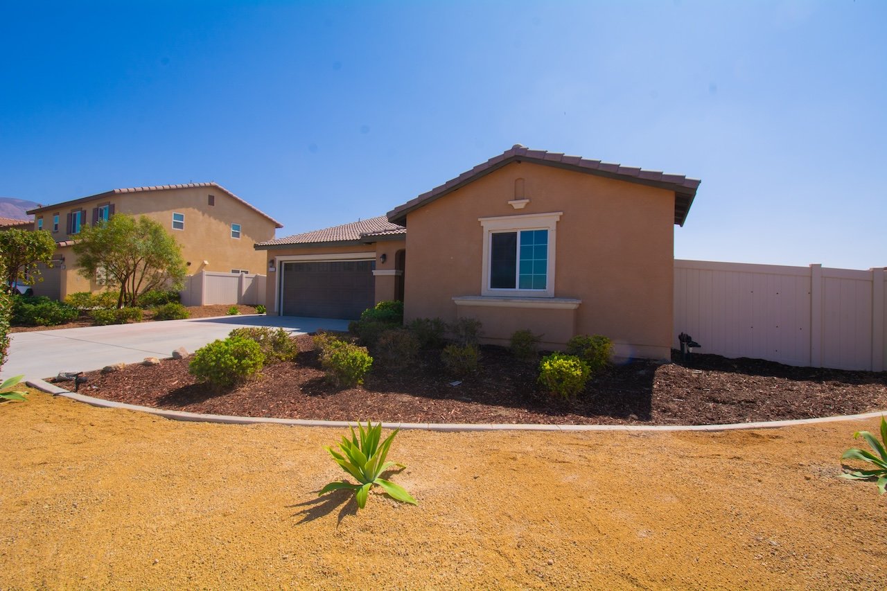 Tan stucco home with an attached garage, pale perimeter fence, and mulch landscaping.