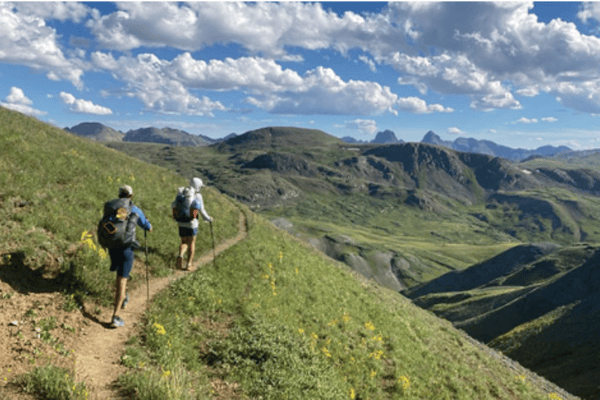 Two hikers follow a trail along a green mountain ridge above a deep valley.