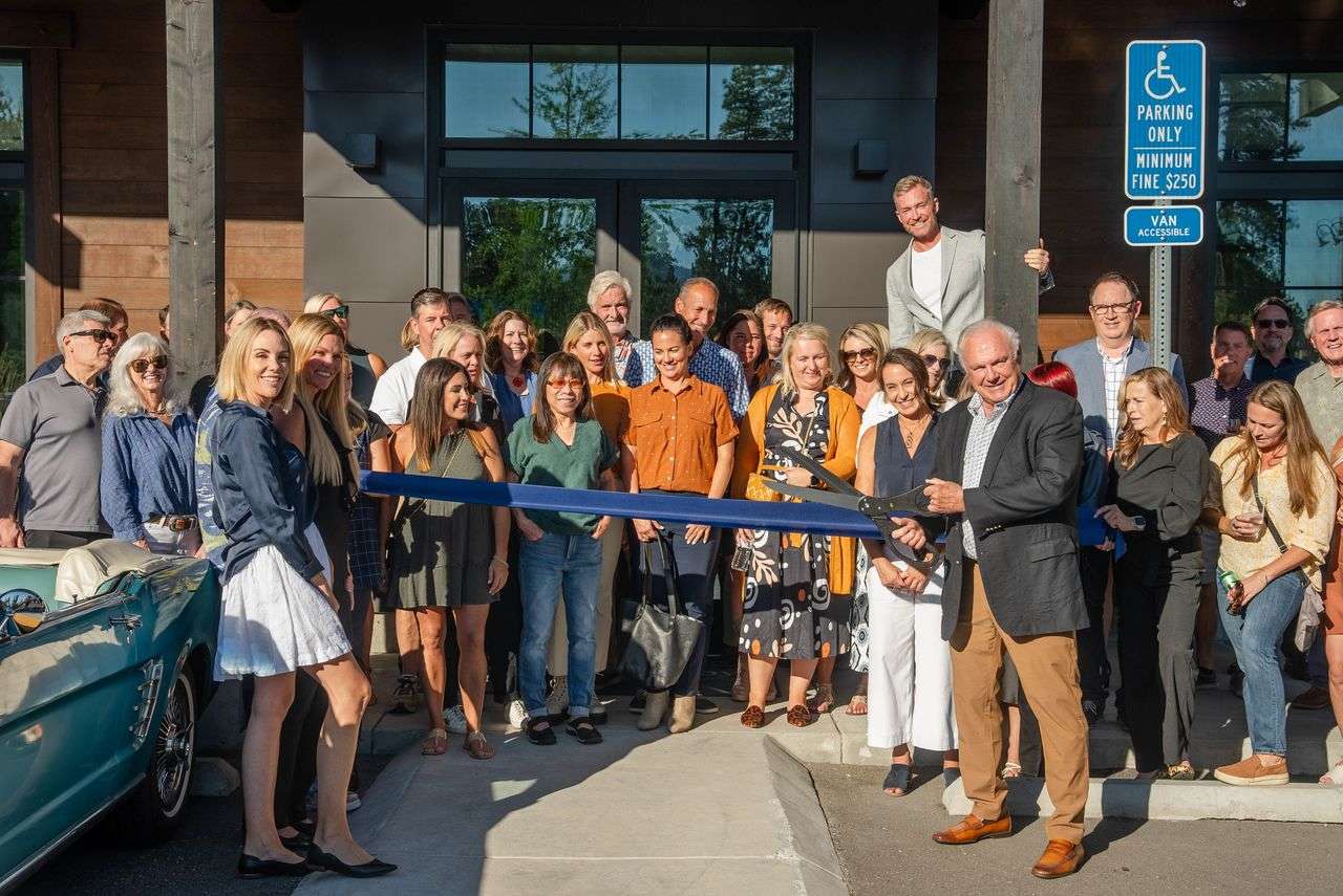 A group poses at an outdoor ribbon cutting, with a blue ribbon and oversized scissors.