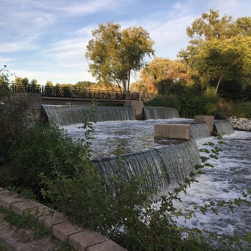 Stepped spillway with rushing water beneath a pedestrian bridge, bordered by autumn trees.