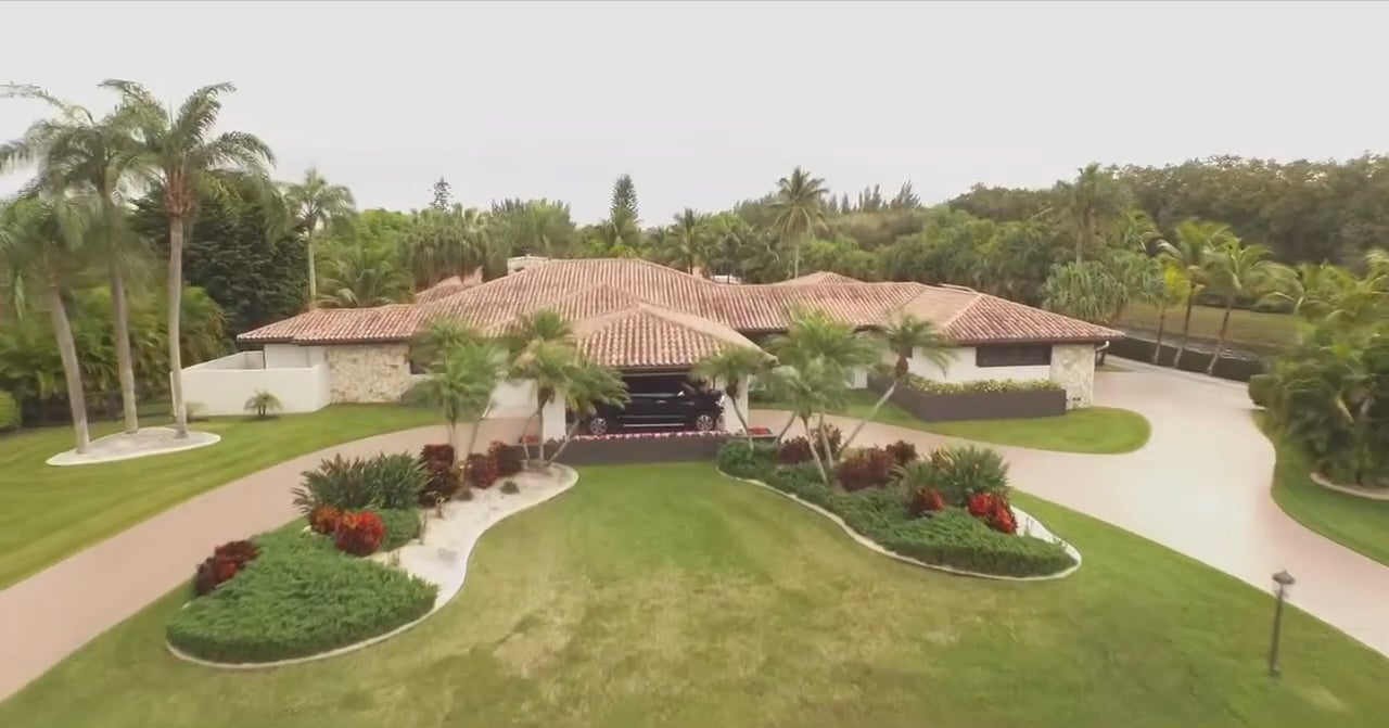 Aerial view of a stone estate with a red tile roof, palm-lined lawn, and curved driveway.