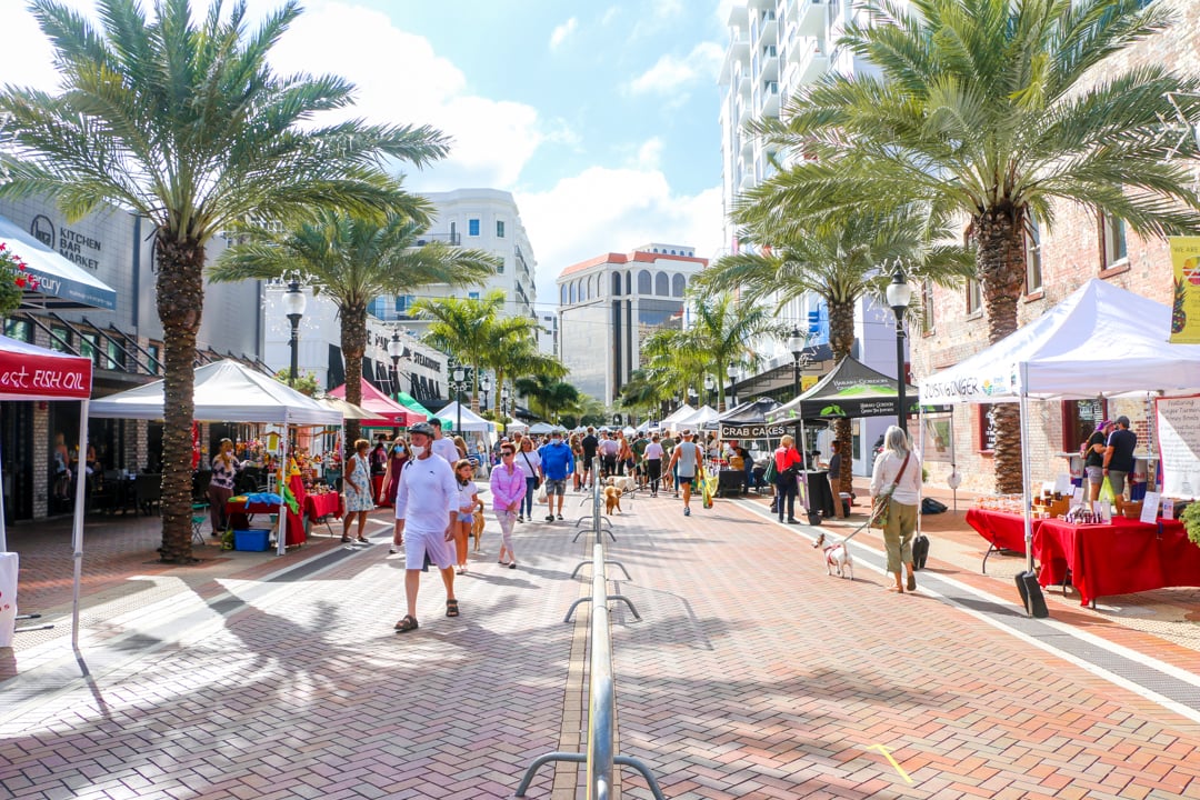 Shoppers browse vendor stalls along a palm-lined, brick-paved farmers market street.