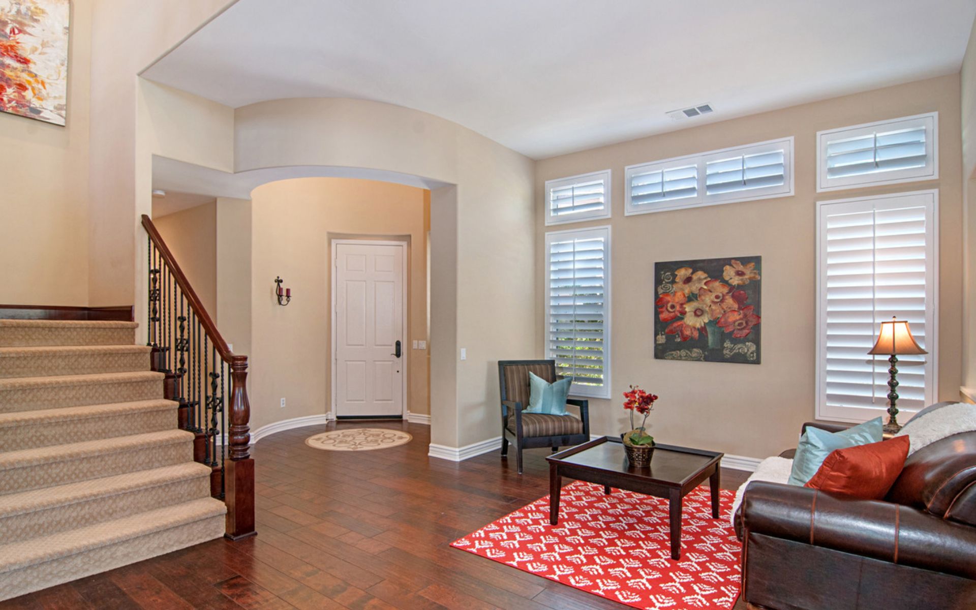 Living room and entryway with a carpeted staircase, shuttered windows, and a red patterned rug.