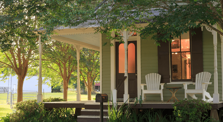 Green house with a covered porch, white Adirondack chairs, and mature trees shading the yard.