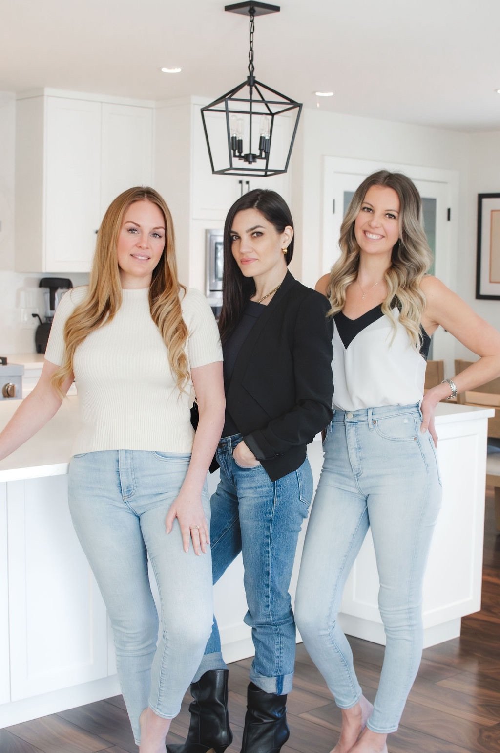 Three women pose in a bright kitchen, one in a black blazer between two women in light tops.