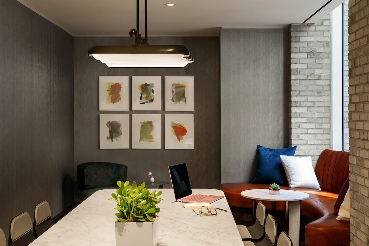 Dining area with a marble table, abstract wall art, and rust-colored banquette beside tall windows.