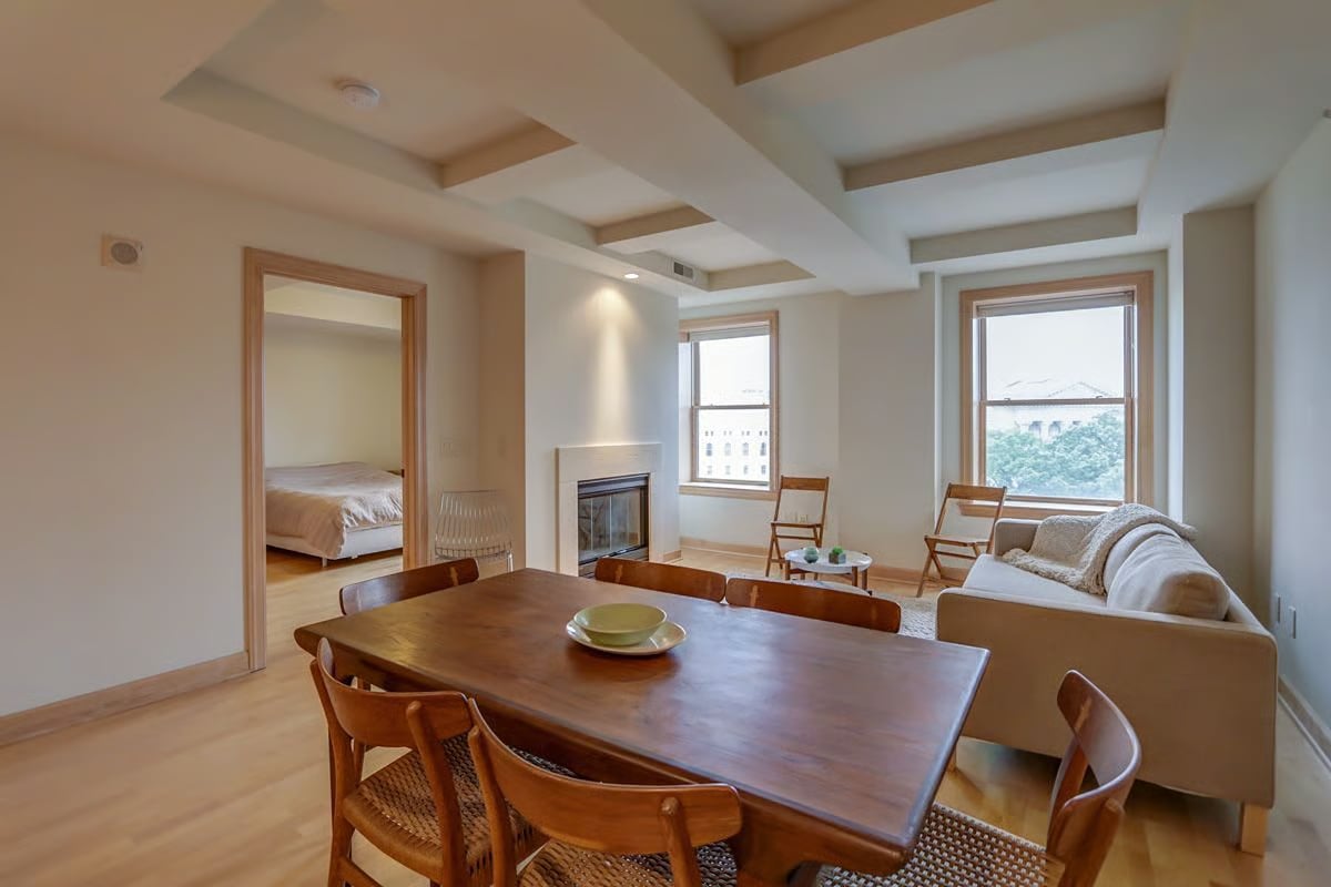 Open-plan living and dining room with a fireplace and windows overlooking greenery and nearby buildings.