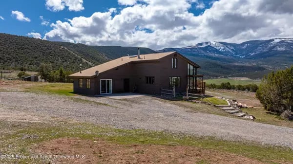 Brown home with a gravel driveway and snow-capped mountains in the distance.