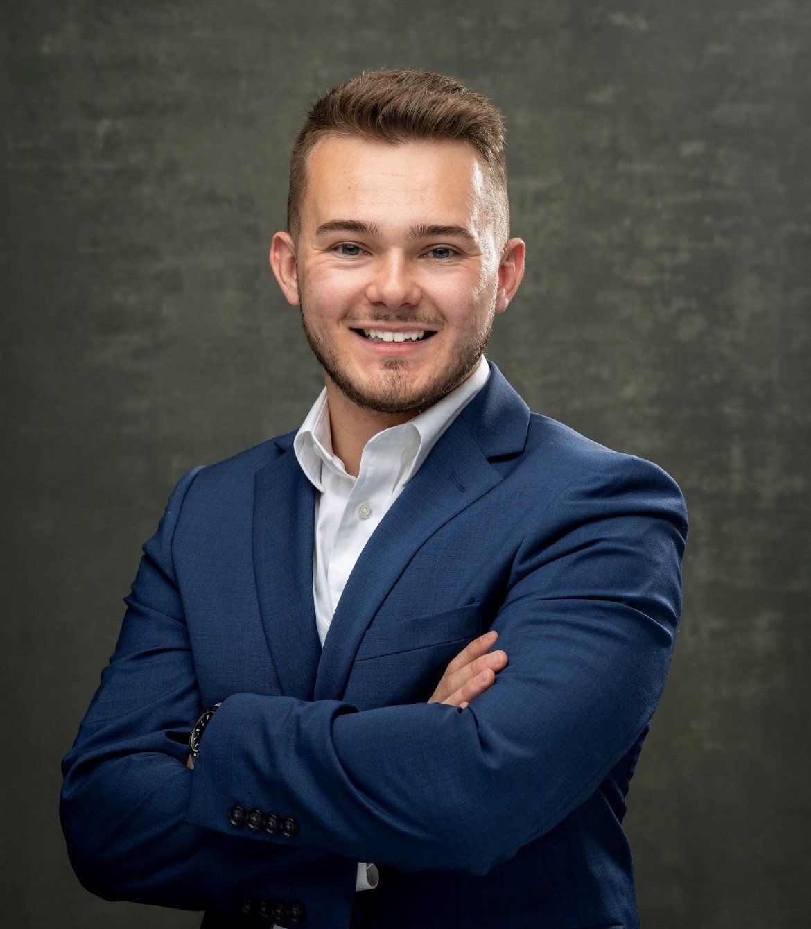 Justin Tewell smiling in a navy suit with his arms crossed.