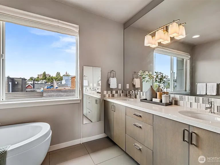 Bathroom with a soaking tub, long double-sink vanity, and large window overlooking rooftops.