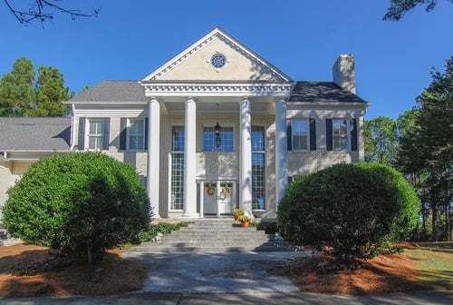 Two-story home with a columned entrance, triangular pediment, and rounded shrubs along the front walkway.