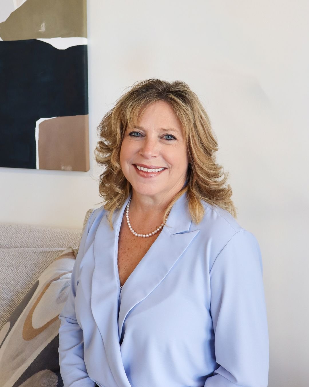 Diane Devries in a pale blue blazer and pearl necklace, seated beside abstract wall art.