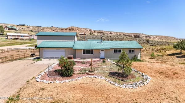 Single-story home with a teal metal roof, attached garage, gravel drive, and stone-edged front landscaping.
