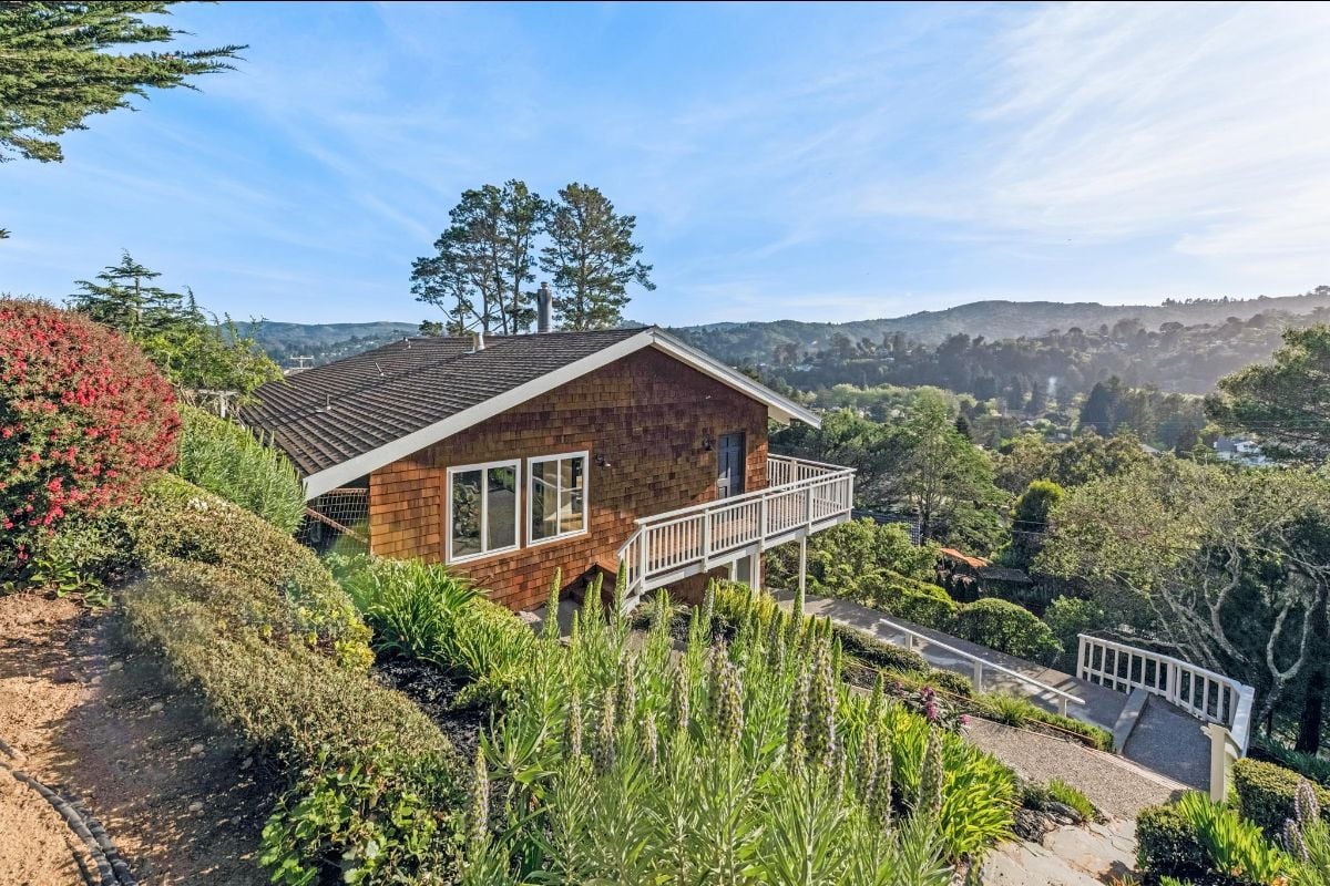 Hillside home with a white-railed deck, landscaped garden, and wooded hills beyond.