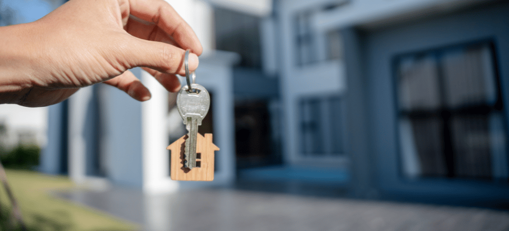 hand holding a keychain with a house key and toy house keychain in front of a modern home