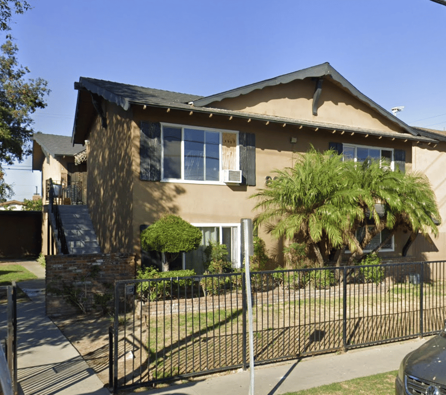 Two-story tan property with dark shutters, palm trees, and a fenced front garden.