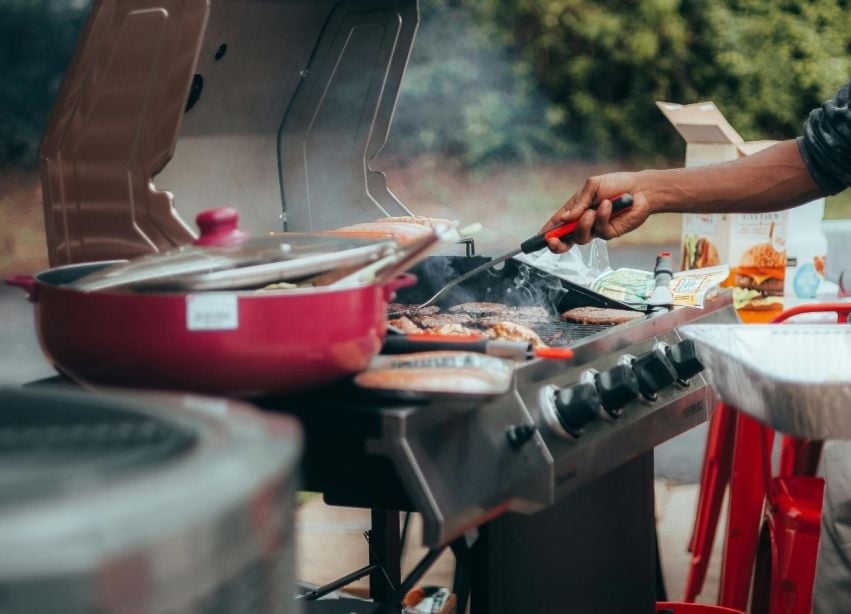 A hand turns food on a smoking outdoor grill beside barbecue supplies.