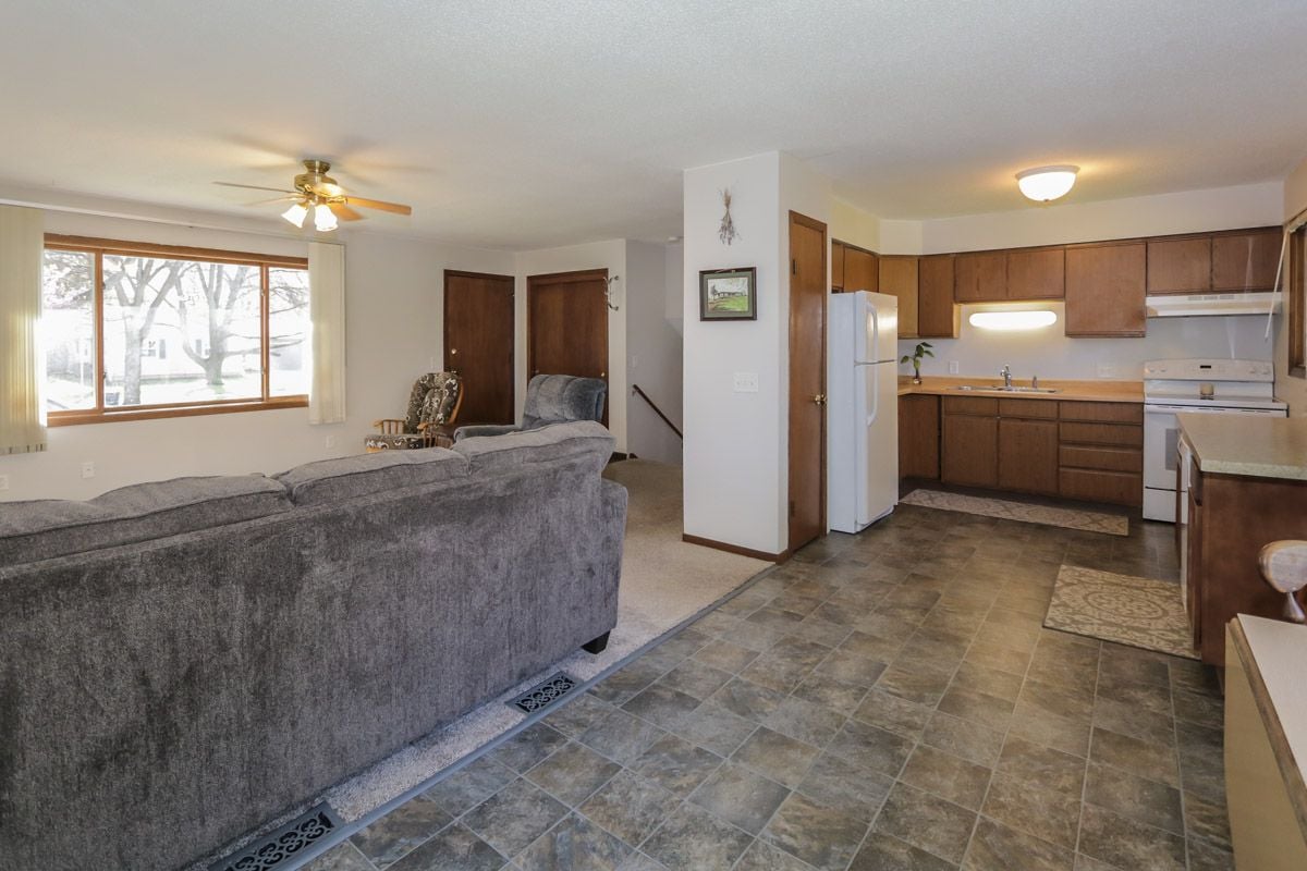 Open-plan living room and kitchen with a gray sofa, wood cabinets, and a large window.