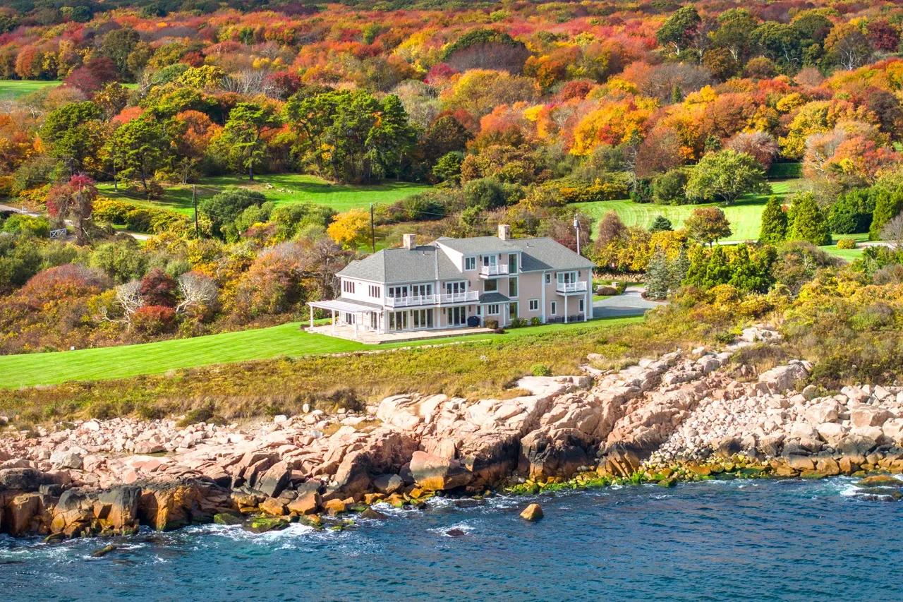 Coastal home above a rocky shoreline, backed by colorful autumn trees.