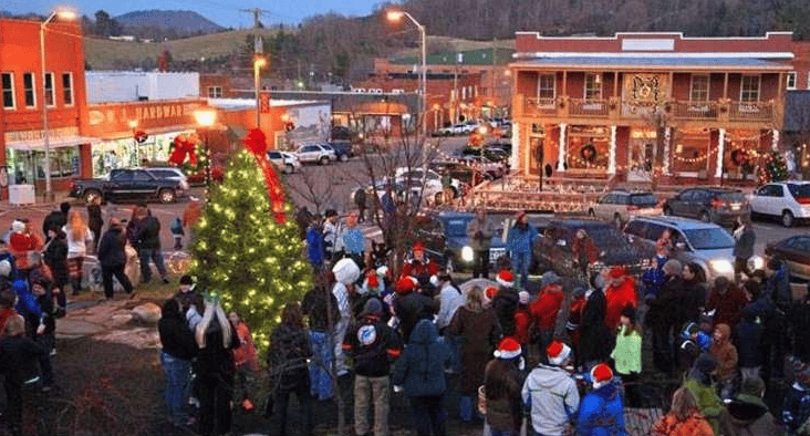 Crowd gathers around a decorated Christmas tree on a downtown street at dusk.