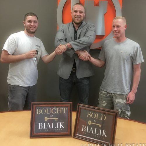 Three men celebrate a real estate closing with handshakes and framed Bought and Sold signs.