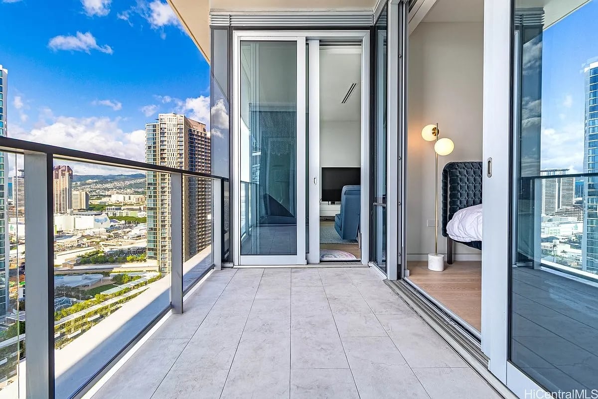 High-rise balcony overlooking the city, with glass railings and sliding doors to a bedroom.
