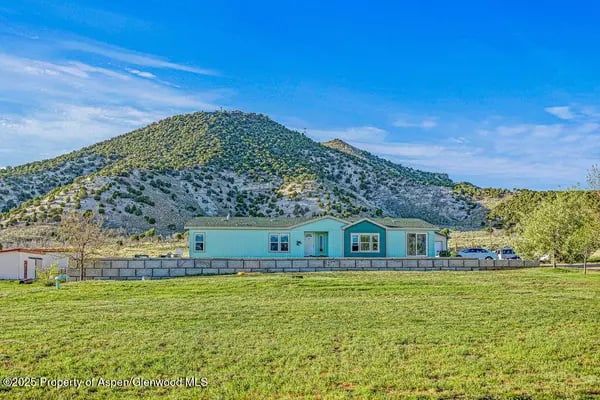 Pale blue single-story home behind a broad lawn, with a shrub-covered mountain in the background.
