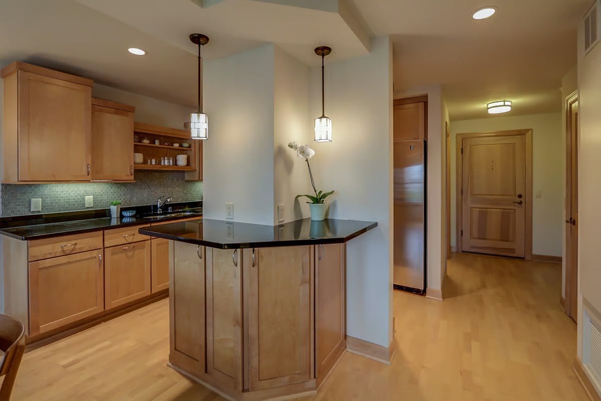 Kitchen with light wood cabinetry, black counters, and a central peninsula beside an entry hallway.